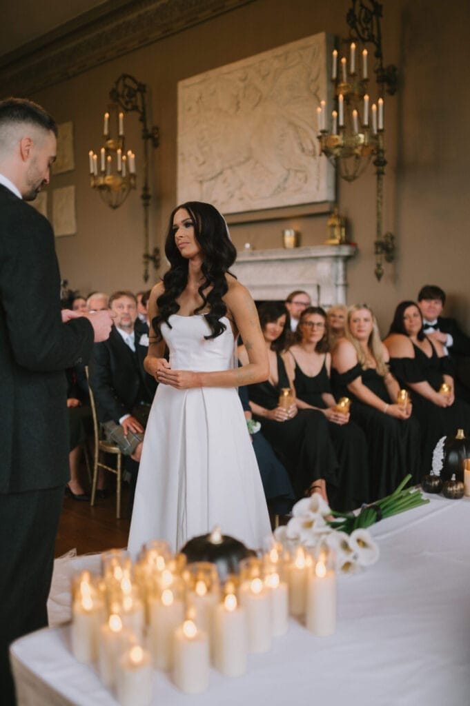 A bride in a white gown stands facing her groom during an indoor ceremony at Howsham Hall. Guests, dressed formally and holding candles, are seated behind her. A table with lit candles is in the foreground—perfect for a Howsham Hall Wedding Photographer to capture.