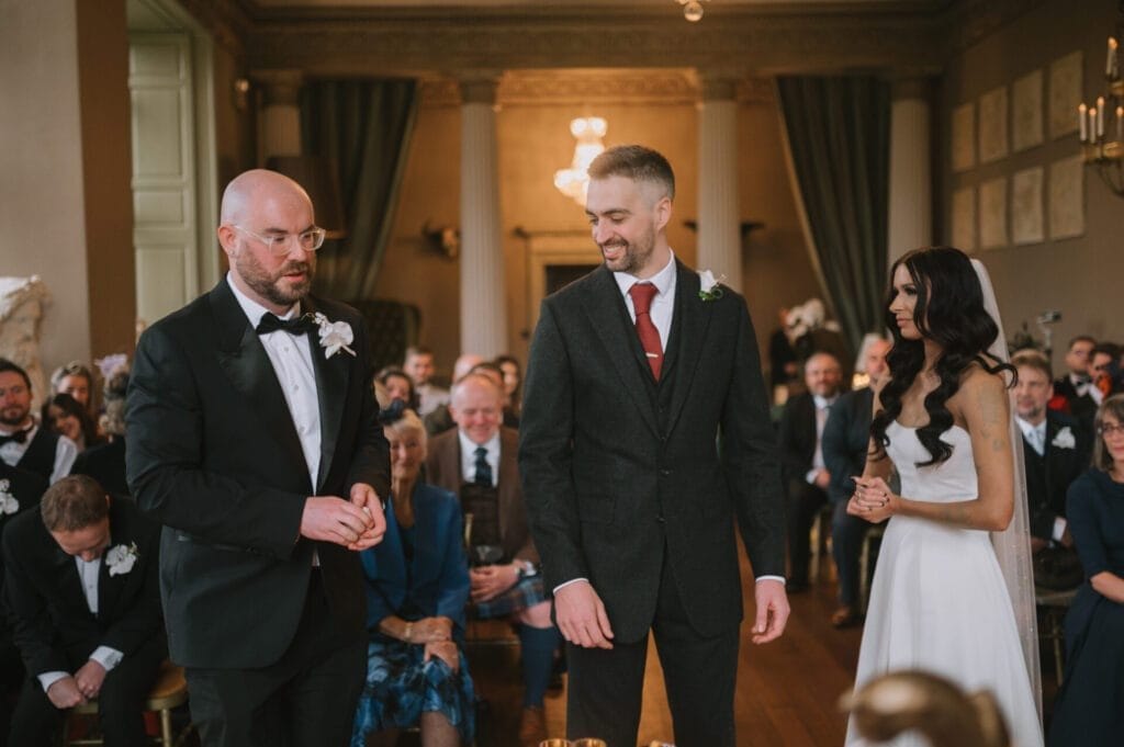 A wedding ceremony at Howsham Hall unfolds in a grand room with chandeliers. A man in a black suit speaks, while another man in gray and a bride stand beside him. Guests watch attentively—captured perfectly by a Howsham Hall Wedding Photographer.