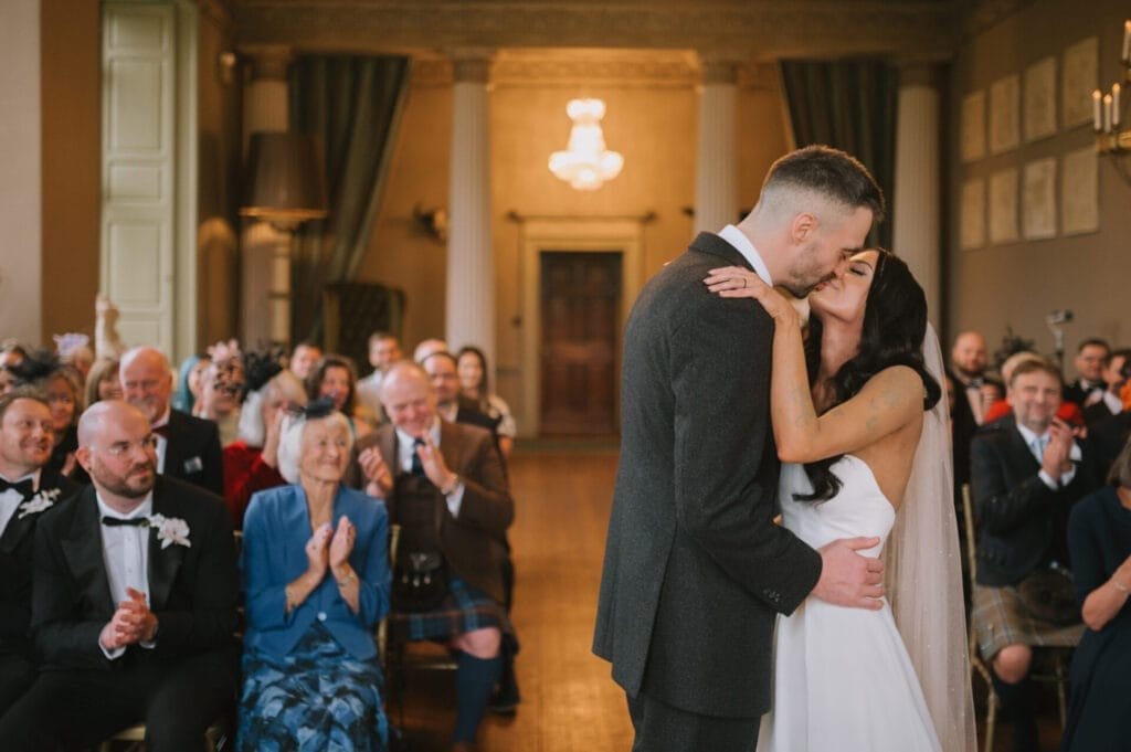 A bride and groom kiss at the altar in a grand room during their wedding ceremony as guests seated around them smile, clap, and watch with joy—captured beautifully by a Howsham Hall Wedding Photographer.