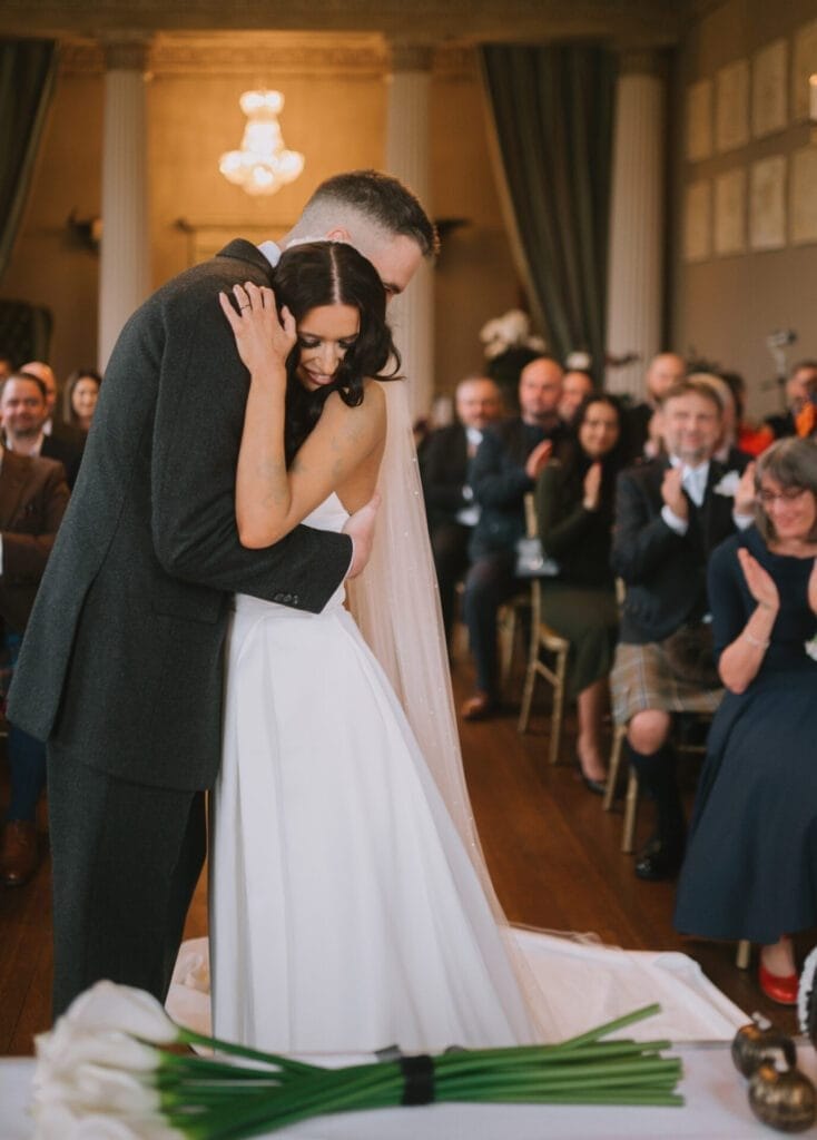 A bride and groom embrace warmly at their wedding ceremony, surrounded by smiling guests. The elegant room, beautifully captured by a Howsham Hall Wedding Photographer, features tall columns, chandeliers, and white flowers in the foreground.