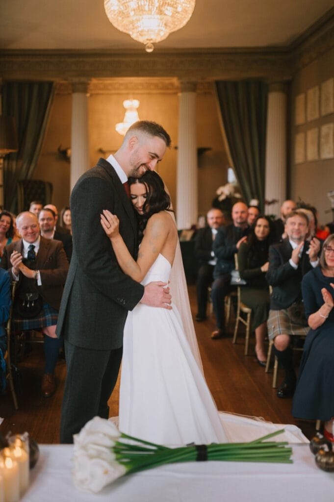 A bride and groom embrace and smile during their wedding ceremony at Howsham Hall, surrounded by seated guests who are watching and applauding. A bouquet of white calla lilies rests on a table in the foreground—a perfect moment for a Howsham Hall Wedding Photographer.