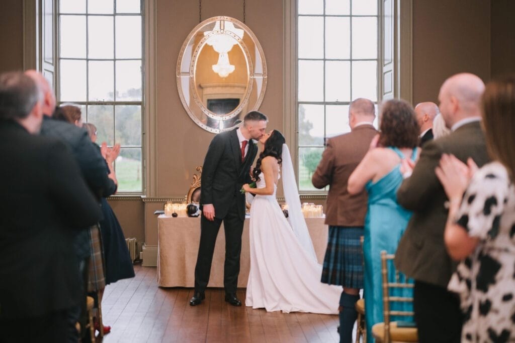 A bride and groom share a kiss at the altar during their indoor wedding ceremony as guests stand, clap, and celebrate their union. Captured by a Howsham Hall Wedding Photographer, a large ornate mirror and tall windows frame the joyful moment.