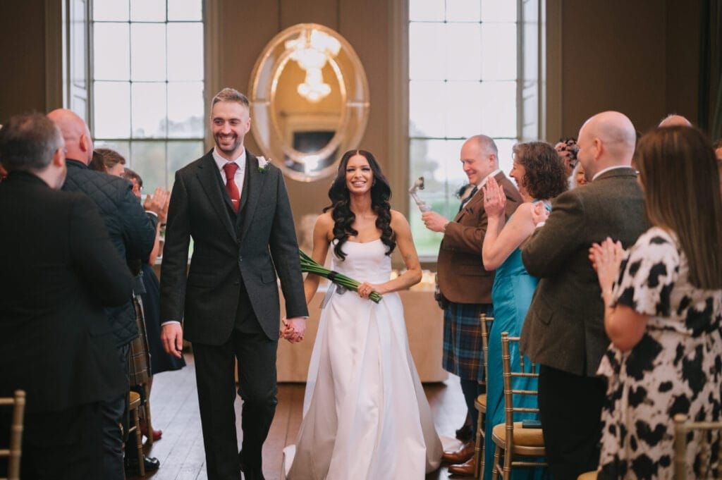 A newlywed couple walks down the aisle, smiling and holding hands, while guests on either side clap and celebrate their marriage in a bright, elegant room captured by a Howsham Hall Wedding Photographer.