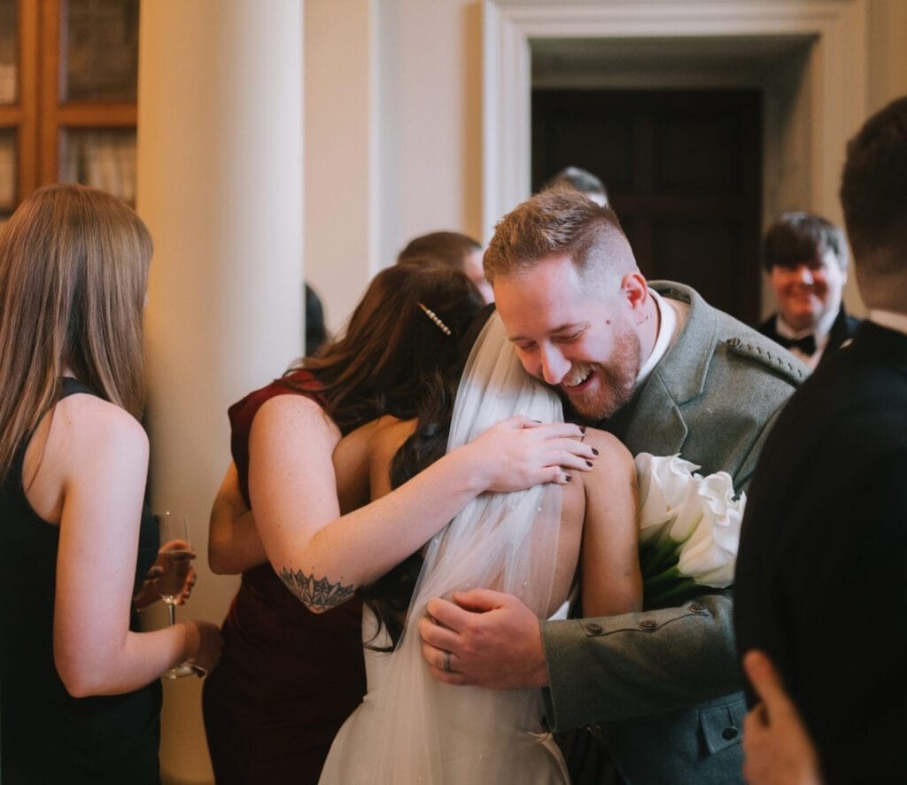 A bride in a white dress and veil hugs a smiling man in a gray suit at an indoor gathering, beautifully captured by a Howsham Hall Wedding Photographer, surrounded by elegantly dressed guests.