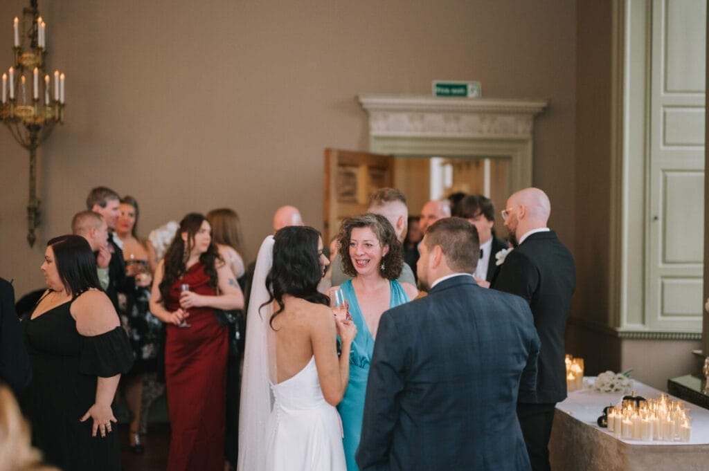 A group of people in formal attire gather indoors at a social event. A woman in a blue dress smiles and talks to a woman in a white dress, while guests mingle and hold drinks near candlelit tables—perfect inspiration for any Howsham Hall Wedding Photographer.