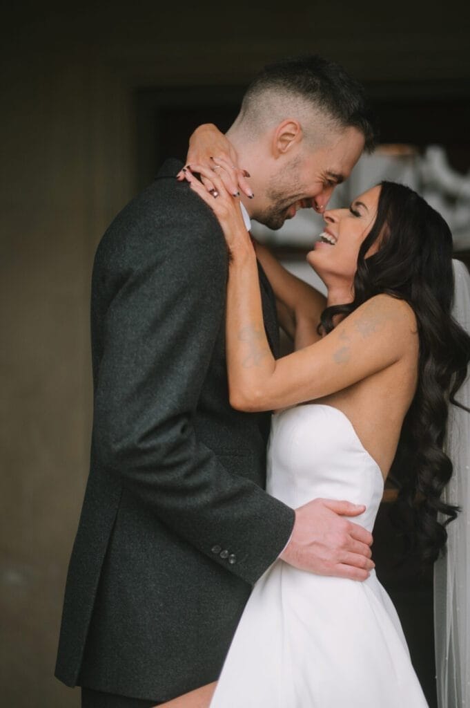 A bride and groom embrace and smile at each other joyfully, standing close together. Captured by a Howsham Hall Wedding Photographer, the happy couple radiate love—the bride in a strapless white dress and the groom in a dark suit.