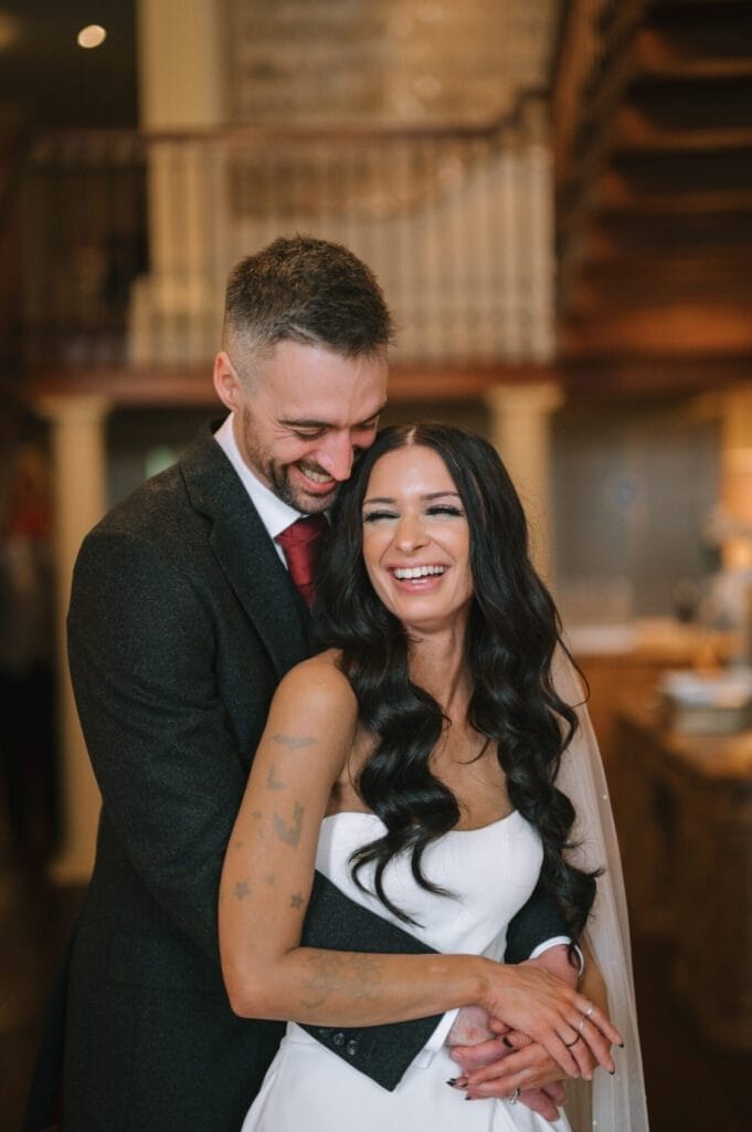 A bride in a white dress and a groom in a suit embrace and laugh together indoors at Howsham Hall, with wooden stairs and railings in the background—captured candidly by a Howsham Hall Wedding Photographer as they share a joyful moment.