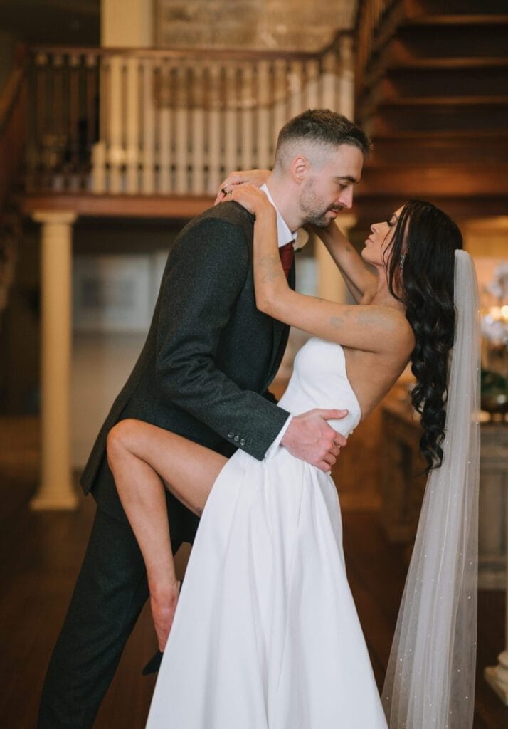 A bride in a white strapless gown and veil playfully lifts her leg as she embraces and smiles at the groom in a dark suit. Captured by a Howsham Hall Wedding Photographer, rustic stairs and railings add charm to their joyful moment indoors.