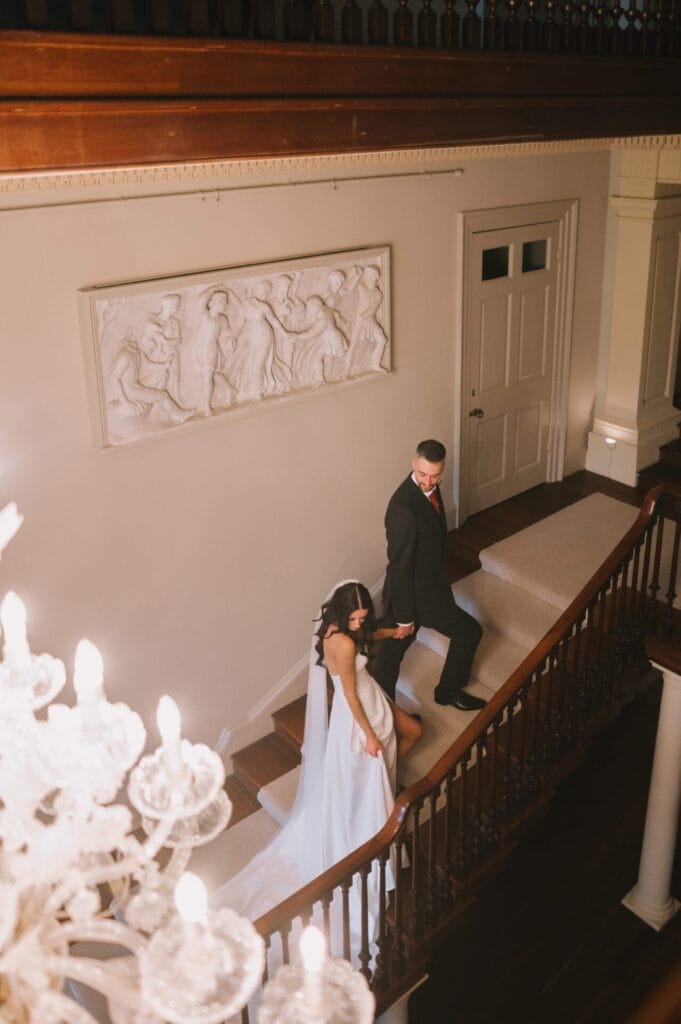 A bride and groom hold hands as they walk up a grand staircase in an elegant, well-lit venue with a chandelier and classical wall art—perfectly captured by a skilled Howsham Hall Wedding Photographer.