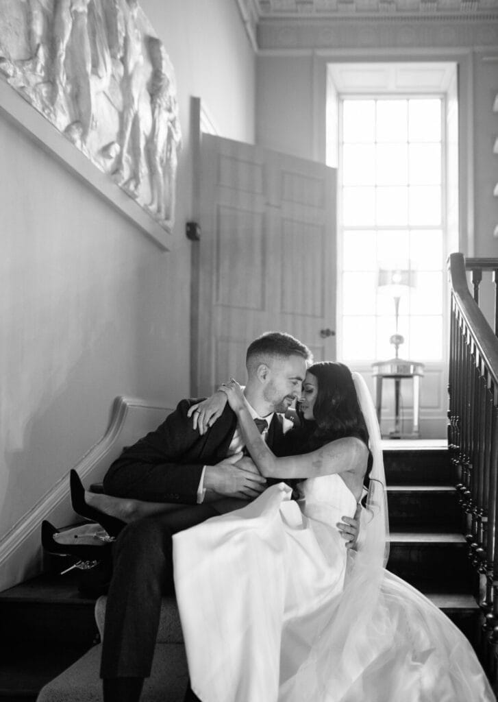 A joyful bride and groom sit on a staircase at Howsham Hall, embracing and smiling at each other. The bride wears a white dress and veil, while the groom is in a suit. Light streams in from a large window behind them.