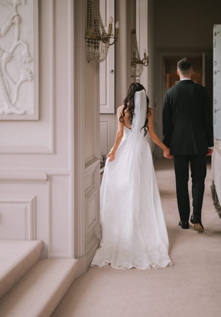 A bride in a white gown and veil holds hands with a groom in a dark suit as they walk down a softly lit hallway at an elegant venue, beautifully captured by a Howsham Hall Wedding Photographer, seen from behind.