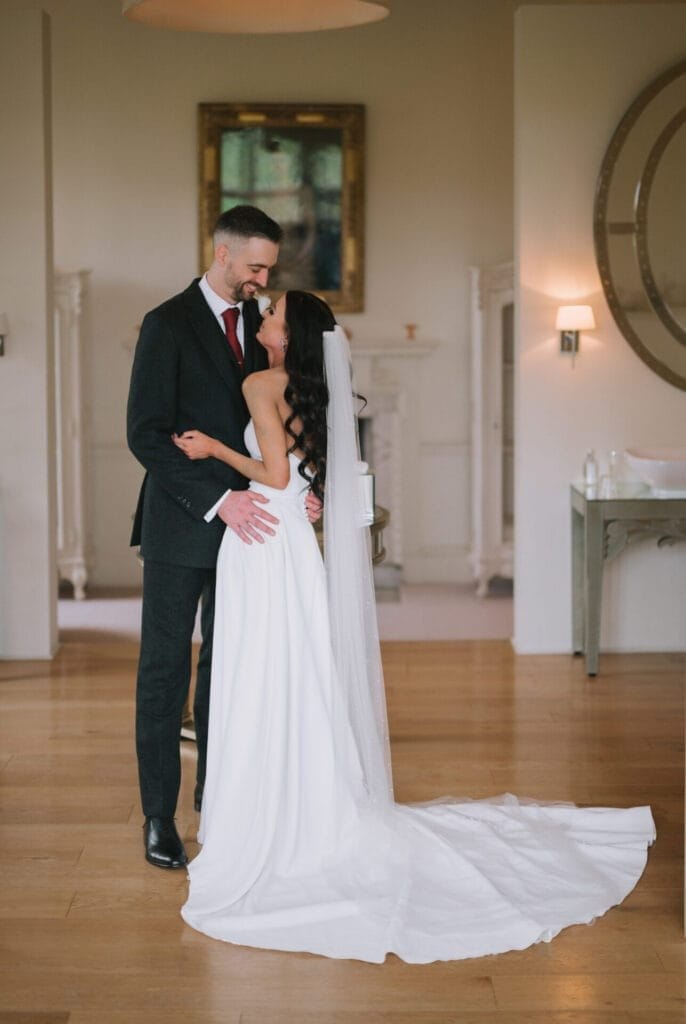 A bride in a white gown and veil stands close to a groom in a dark suit and red tie, gazing at each other and smiling in an elegant indoor setting with wooden floors and soft lighting, perfectly captured by a Howsham Hall Wedding Photographer.