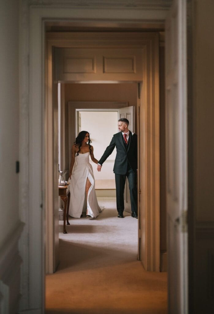 A bride in a white dress and a groom in a black suit hold hands and walk through a doorway in an elegant, softly lit hallway, perfectly captured by a Howsham Hall Wedding Photographer.