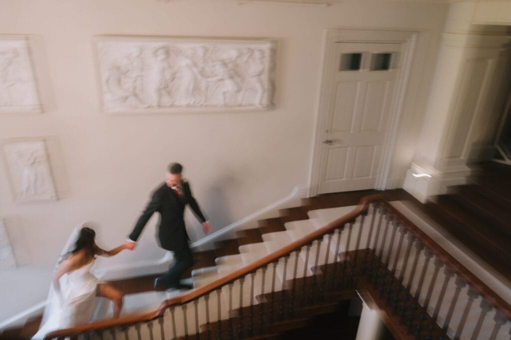 A bride in a white dress and veil holds hands with a groom in a dark suit as they walk up a staircase. Captured by a Howsham Hall Wedding Photographer, the slightly blurred photo reveals classical reliefs on the wall behind them.