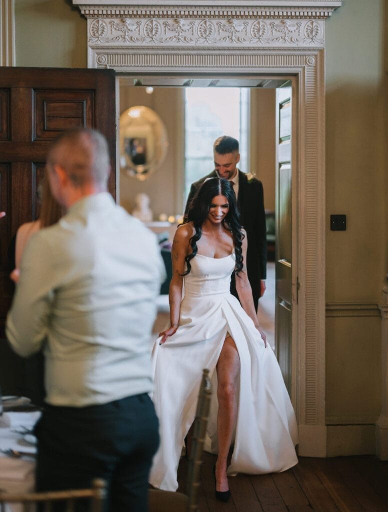 A bride in a white dress with a thigh-high slit walks through a doorway, smiling, with a man in a suit behind her. Guests are seated at tables in an elegant, decorated room captured by a Howsham Hall Wedding Photographer.