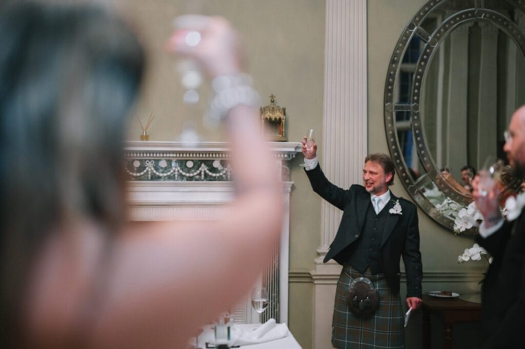 A man in a kilt raises a glass for a toast in an elegant room with a large round mirror, while others join in. Capturing this joyful scene, the Howsham Hall Wedding Photographer highlights the celebratory spirit of the event.