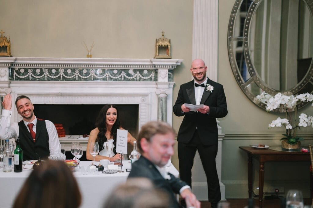 A man in a tuxedo gives a speech at a wedding reception as the bride and groom laugh. The elegant room, perfect for a Howsham Hall Wedding Photographer, features a large round mirror and an ornate fireplace.