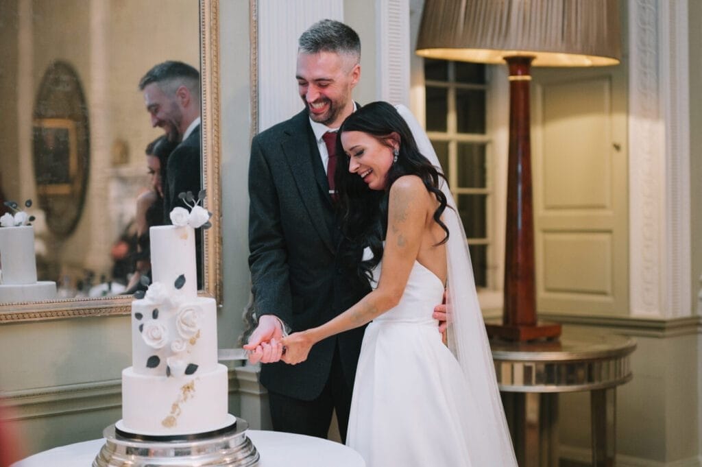A bride and groom smile as they cut a white, cow-print wedding cake together in an elegant room at Howsham Hall, with a large mirror, lamp, and classic decor—captured perfectly by their wedding photographer.