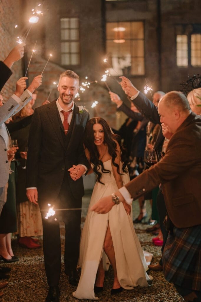 A bride and groom walk hand in hand, smiling joyfully, as guests hold sparklers around them at night. Captured by a Howsham Hall Wedding Photographer, the couple is surrounded by friends and family celebrating outdoors.