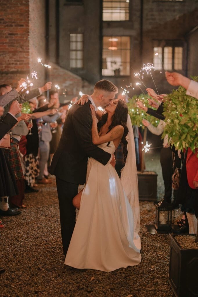 A bride and groom kiss passionately outdoors at night, surrounded by guests holding sparklers. Captured by a Howsham Hall Wedding Photographer, this joyful and romantic celebration showcases the bride in white and the groom in a dark suit.