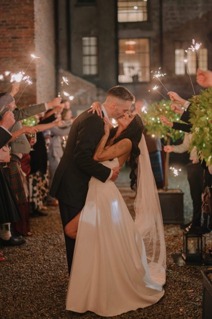 A bride and groom share a kiss during an outdoor evening celebration at Howsham Hall, surrounded by guests holding sparklers. The bride wears a white dress and veil, the groom is in a suit, with warm lights and greenery creating a festive atmosphere.
