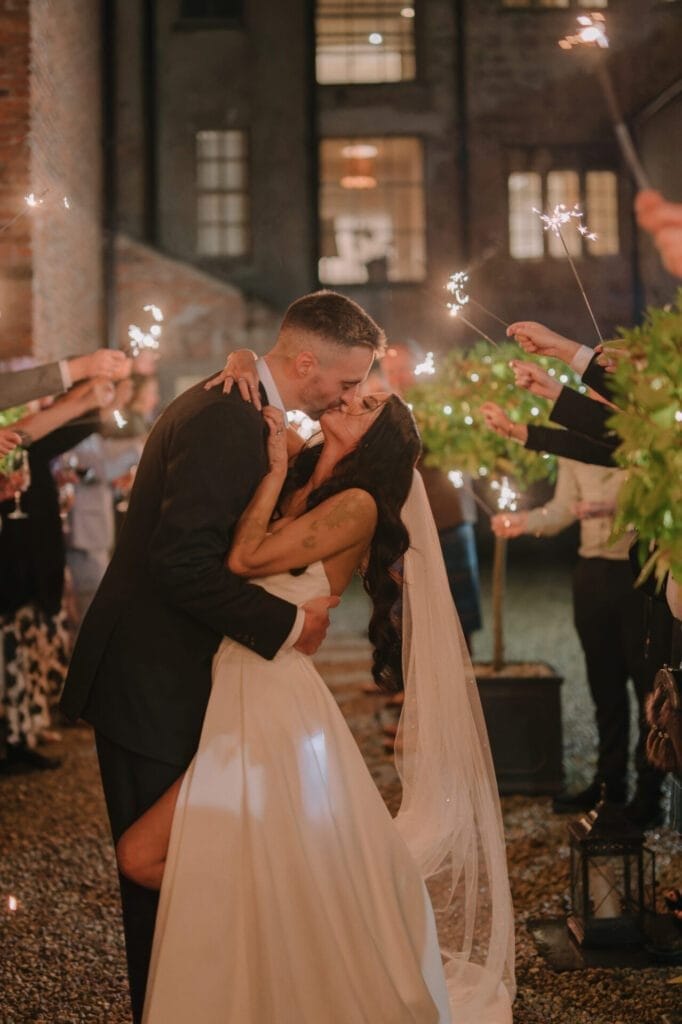 A bride and groom share a passionate kiss outside at night, surrounded by guests holding sparklers. Captured by a Howsham Hall Wedding Photographer, the bride wears a white gown and veil; the groom is in a black suit as warm lights glow behind them.