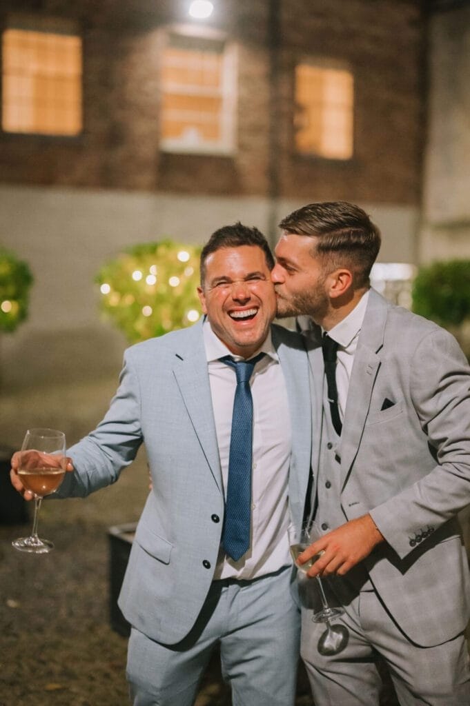 Two men in light-colored suits stand outside at night—one laughs with his eyes closed, wine in hand, while the other smiles, kissing his cheek and holding a drink. Captured beautifully by a Howsham Hall Wedding Photographer.