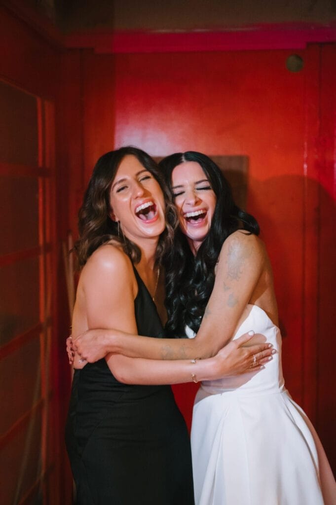 Two women in formal dresses, one in black and one in white, are hugging and laughing joyfully in front of a red wall—captured beautifully by a Howsham Hall Wedding Photographer.
