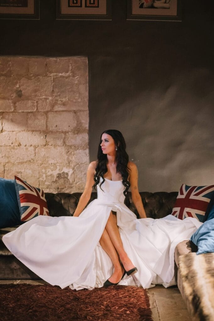 A woman in a white gown with long dark hair sits on a sofa with Union Jack pillows, looking to her left, in a dimly lit room with stone and dark walls, captured by a Howsham Hall Wedding Photographer.
