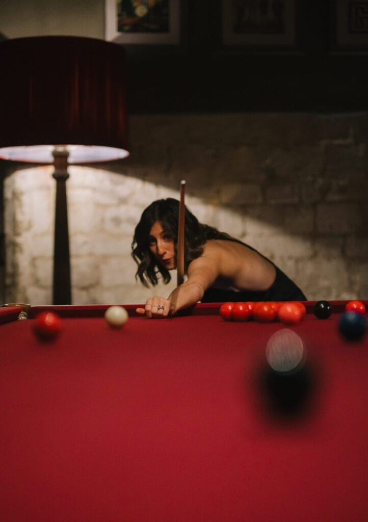 A woman in a black dress leans over a red pool table, aiming her cue at the cue ball. With a lamp glowing and a stone wall behind her, this scene captures the elegance a Howsham Hall Wedding Photographer might seek.