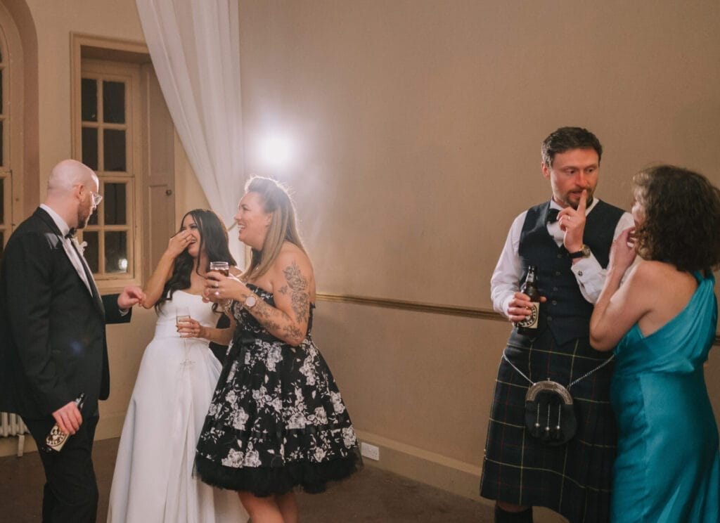 A group of five people dressed formally stand together at a party, laughing and talking. Captured by a Howsham Hall Wedding Photographer, one person is in a wedding dress, another in a kilt, as everyone enjoys drinks and the celebration.