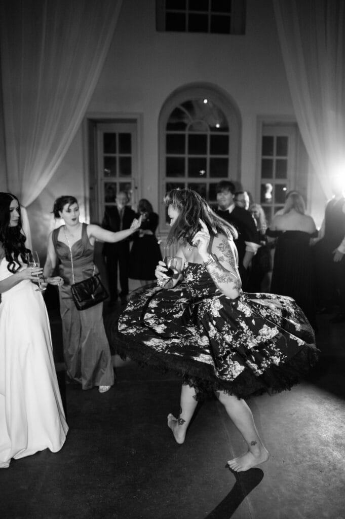 A barefoot woman in a floral dress spins joyfully on the dance floor at a formal event at Howsham Hall, surrounded by guests in evening attire. The lively scene is perfect for any Howsham Hall Wedding Photographer to capture.