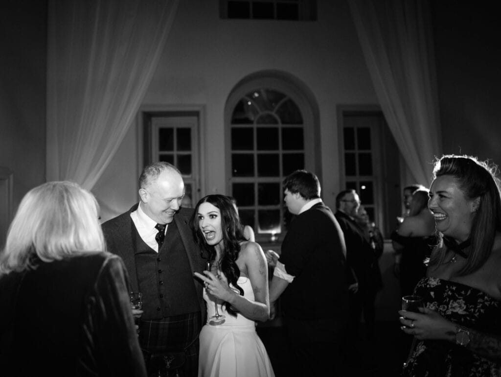 A bride and groom smile and chat with guests at their wedding reception. The softly lit room, featuring large windows and draped curtains, provides a stunning setting captured beautifully by a Howsham Hall Wedding Photographer.