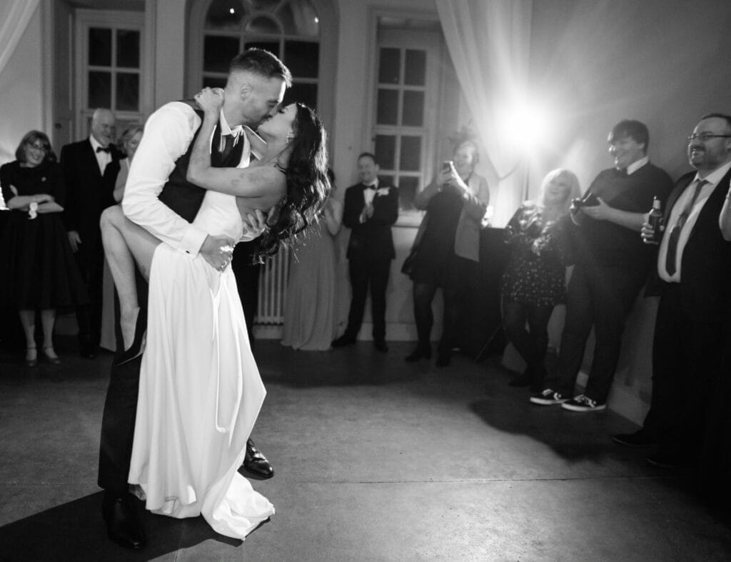 A couple shares a joyful, romantic dance at a wedding reception, surrounded by smiling guests snapping photos. The scene is lit by a bright flash, perfectly captured by a skilled Howsham Hall Wedding Photographer.