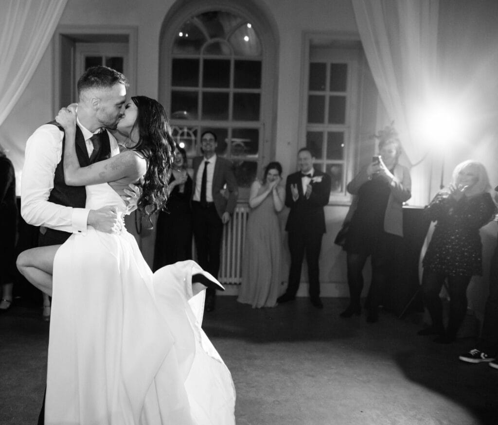 A bride and groom share a passionate kiss while dancing at their wedding, surrounded by cheering guests snapping photos in a warmly lit room—perfect moments captured by a Howsham Hall Wedding Photographer.