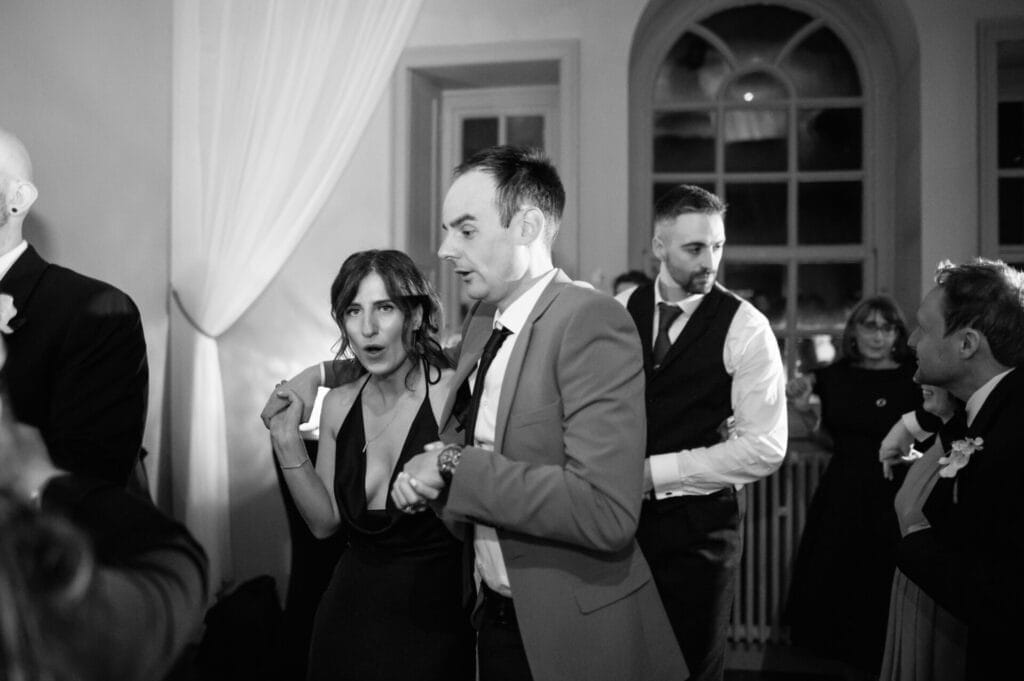 A group of people dressed formally dance together at an indoor event. A woman in a dark dress and a man in a suit are in the foreground, captured by a Howsham Hall Wedding Photographer, while others socialize and dance under soft lighting.