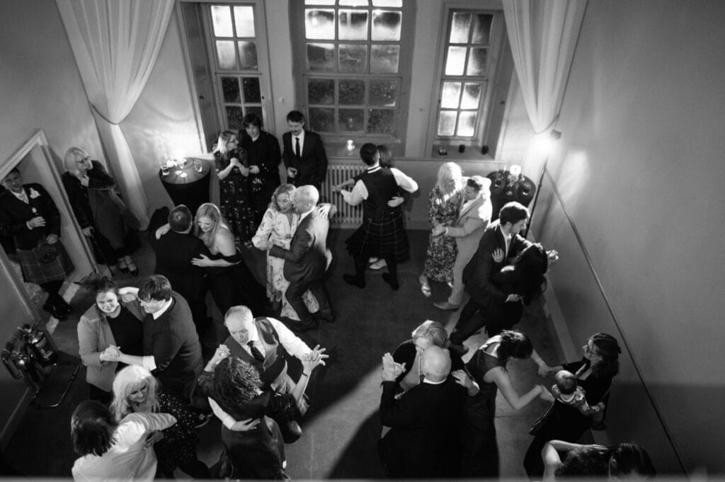 A black and white overhead photo by a Howsham Hall Wedding Photographer shows several couples dancing in a warmly lit room with tall windows and curtains, while others stand nearby, creating a lively, joyful atmosphere.