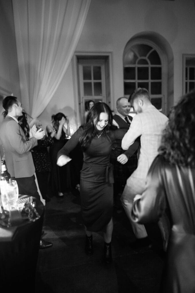 A black and white photo of people dancing and enjoying a lively event in a room with high windows and curtains, captured by a Howsham Hall Wedding Photographer. A woman smiles as she dances with a man in a suit while others clap and watch in the background.