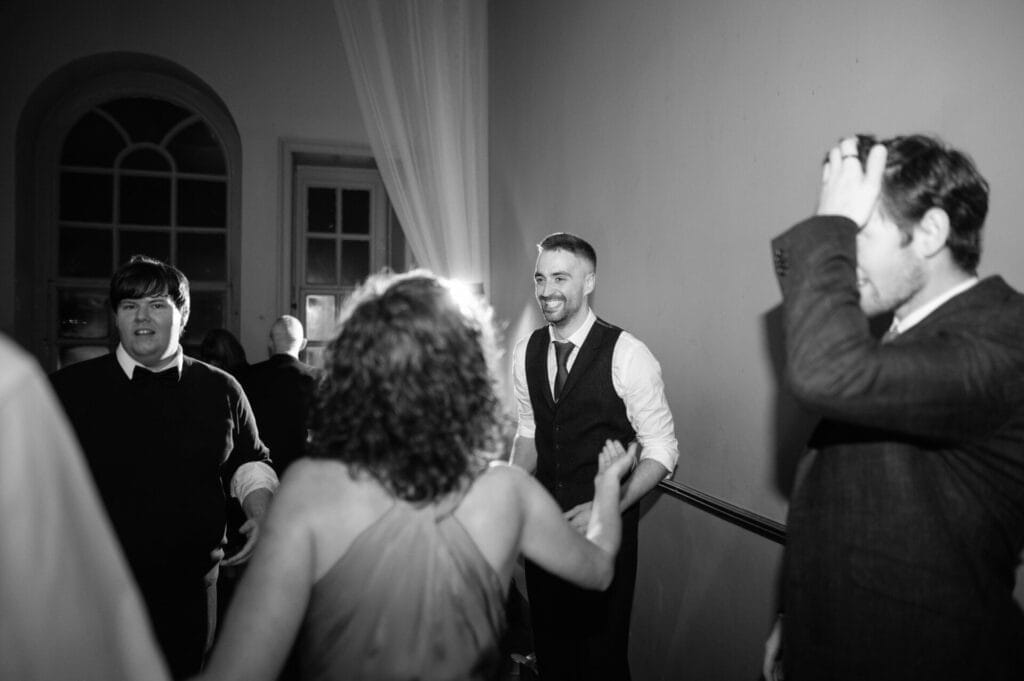 Four adults are gathered in a dimly lit room, smiling and talking. One man in a vest laughs, a woman in a dress has her back to the camera, and another man adjusts his hair. The lively scene captures the spirit of a Howsham Hall Wedding Photographer’s lens.