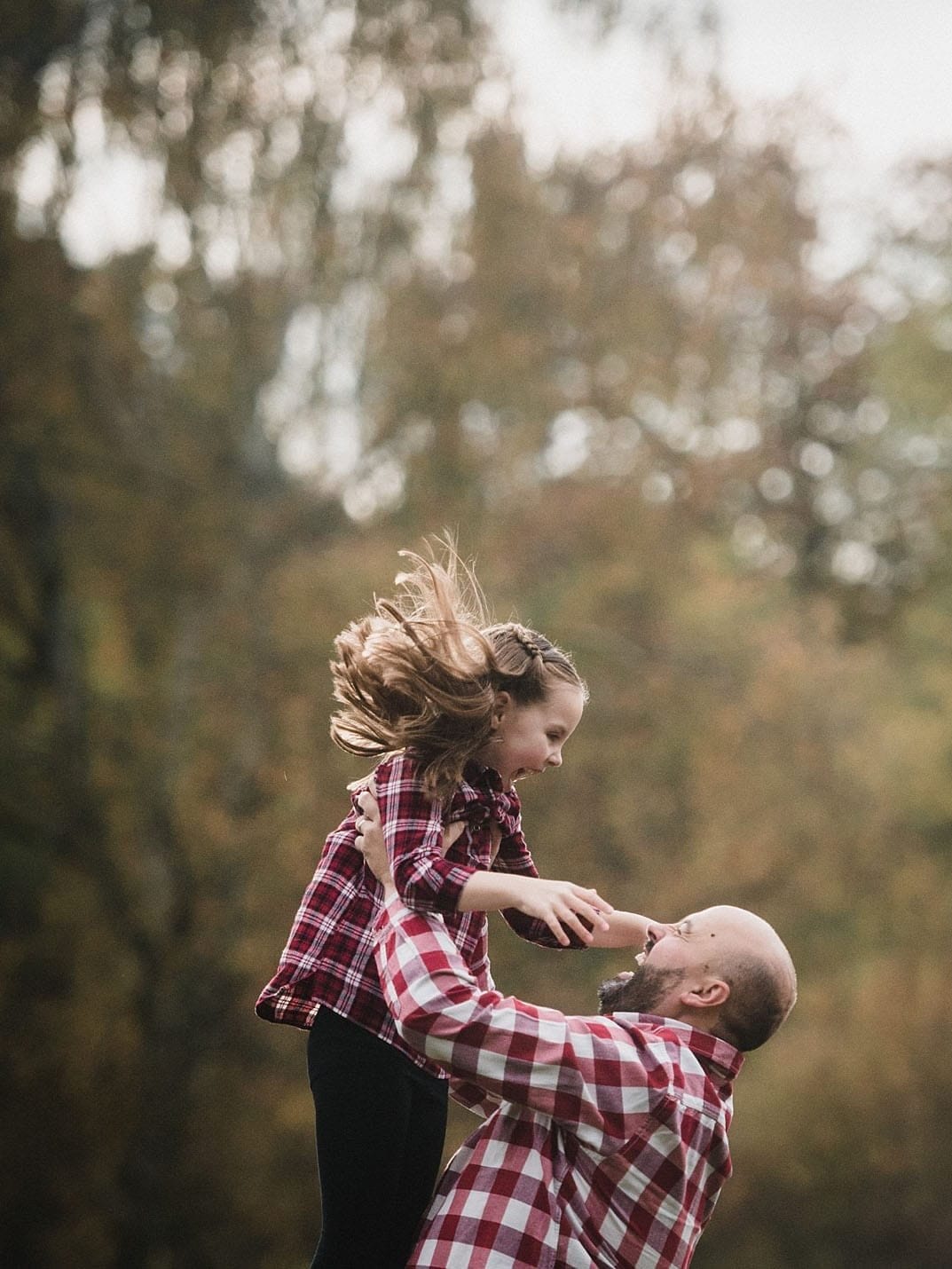A man in a red plaid shirt lifts a laughing young girl, also in a red plaid shirt, into the air outdoors. The background is blurred with autumn trees.