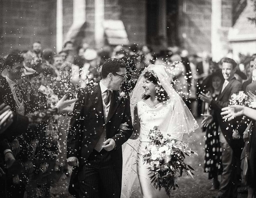 A newlywed couple, smiling and holding hands, walks through a crowd of guests who are throwing confetti. The bride holds a bouquet and wears a veil, while the groom is in a suit. The scene is joyful and celebratory.