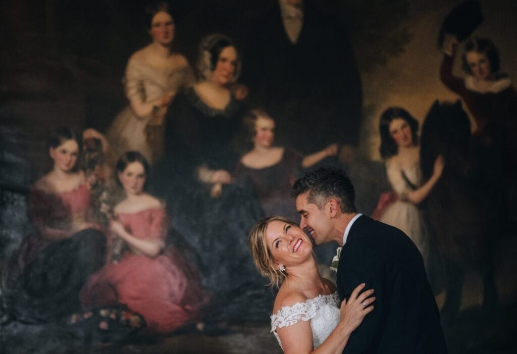 A bride and groom share a joyful embrace captured by a Norwood Park wedding photographer, set against a large, classical painting of women in elegant dresses. The bride, in her white gown, smiles up at the groom in his suit as he leans in closely.