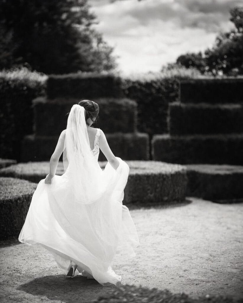 A black and white photo captures a bride at Temple Newsam, walking through a garden maze in a flowing wedding dress. Her back faces the camera, veil cascading down as she gently lifts her gown off the ground. Its a sunny day with fluffy clouds dotting the sky, perfect for wedding photography.
