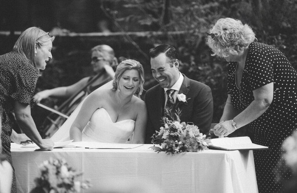 A bride and groom sit at a table, smiling and signing documents, surrounded by two women. A musician playing a cello is in the background. The scene is outdoors and joyful, captured in black and white.