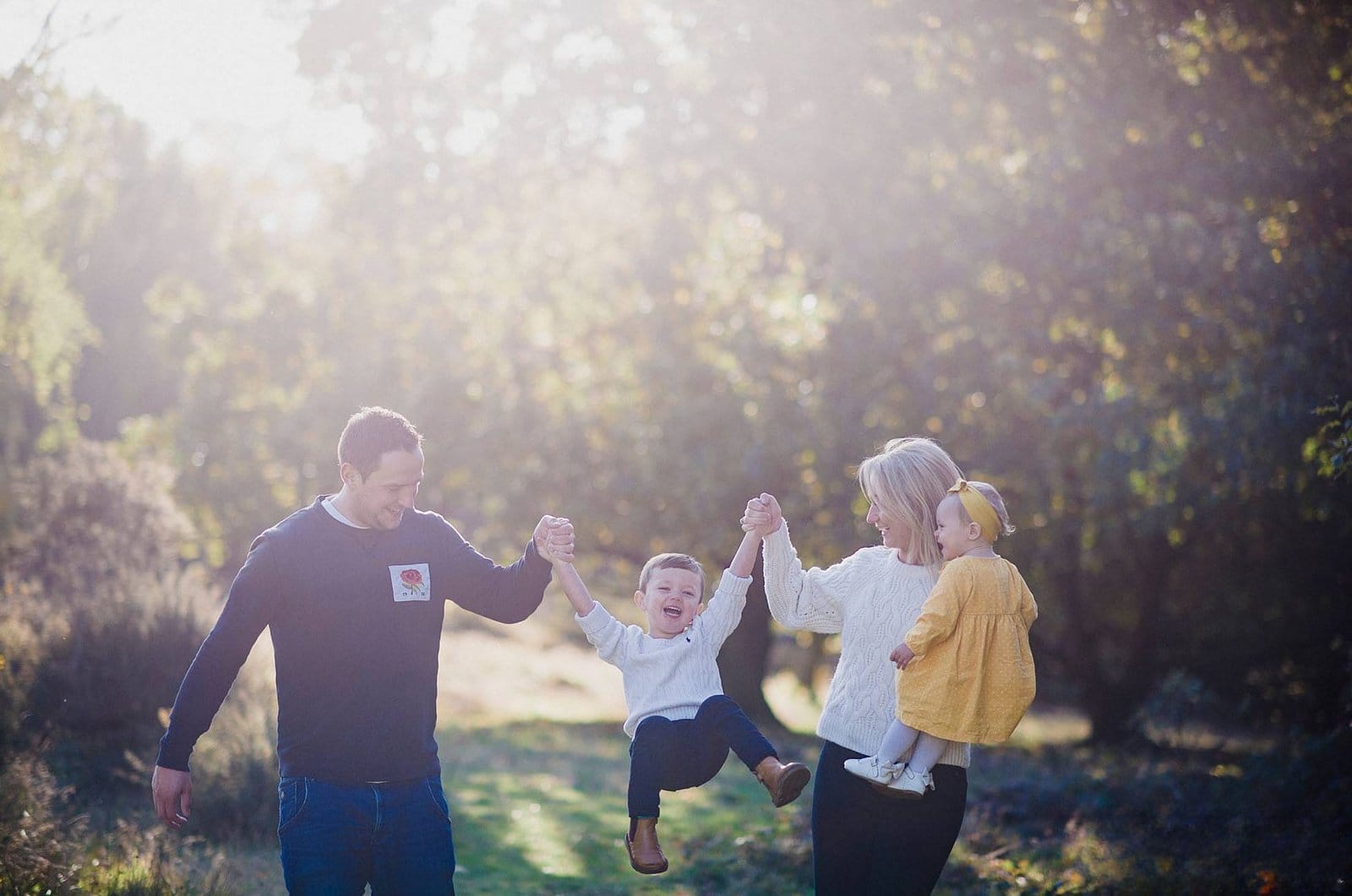A smiling family of four walks outdoors. The father and mother hold hands with a young boy, swinging him between them. The mother carries a toddler girl in a yellow dress. Sunlight filters through the trees in the background.
