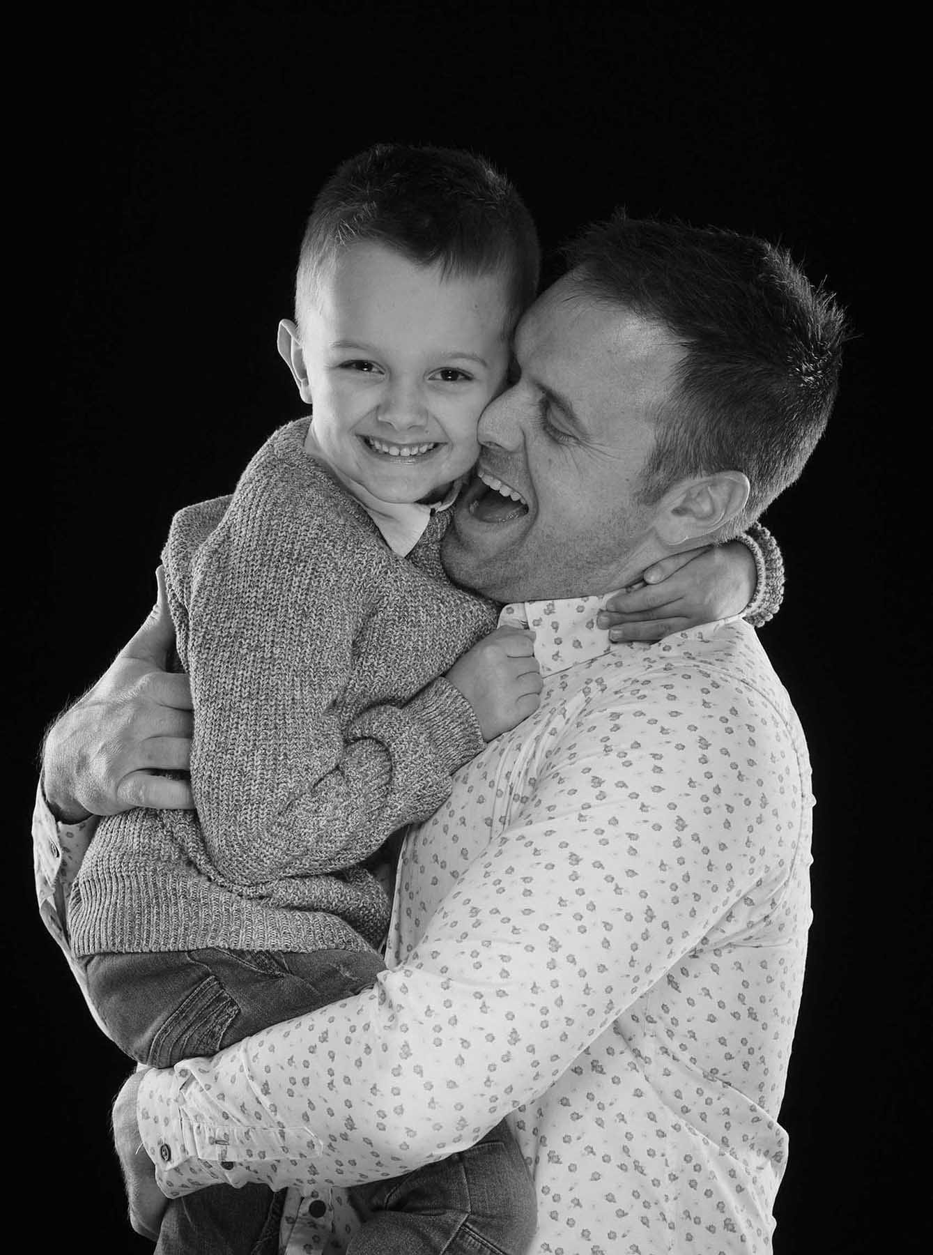 A man smiling and holding a young boy in his arms. Both are happy, and the boy is grinning while wearing a sweater. The background is black, and they are close together in a joyful moment.
