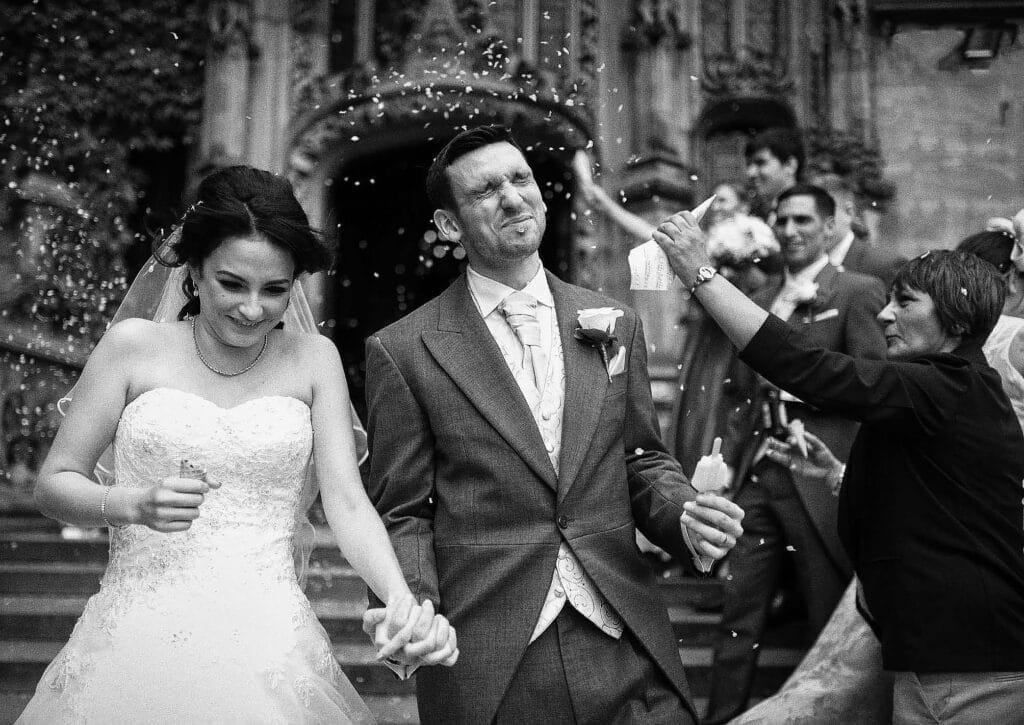 A bride and groom hold hands and smile as they walk outside, while guests throw confetti. The groom squints and cringes as confetti lands on his face, and the bride laughs, surrounded by people celebrating.