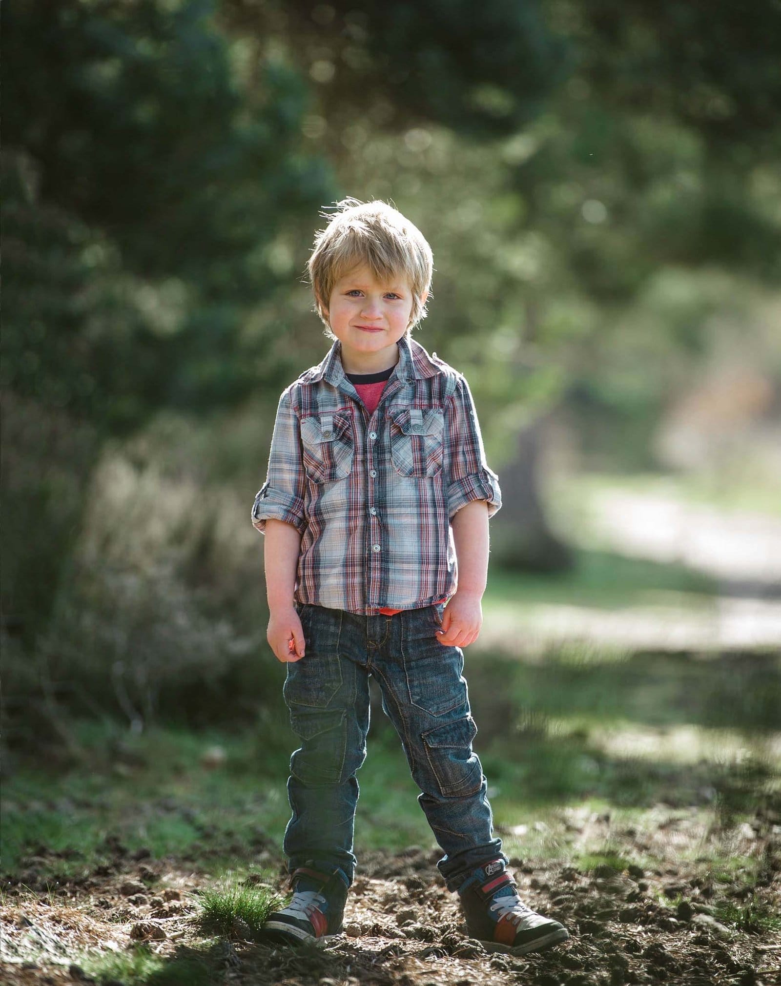A young boy with blonde hair, wearing a plaid shirt, jeans, and sneakers, stands outdoors on a dirt path surrounded by greenery and trees, smiling at the camera.
