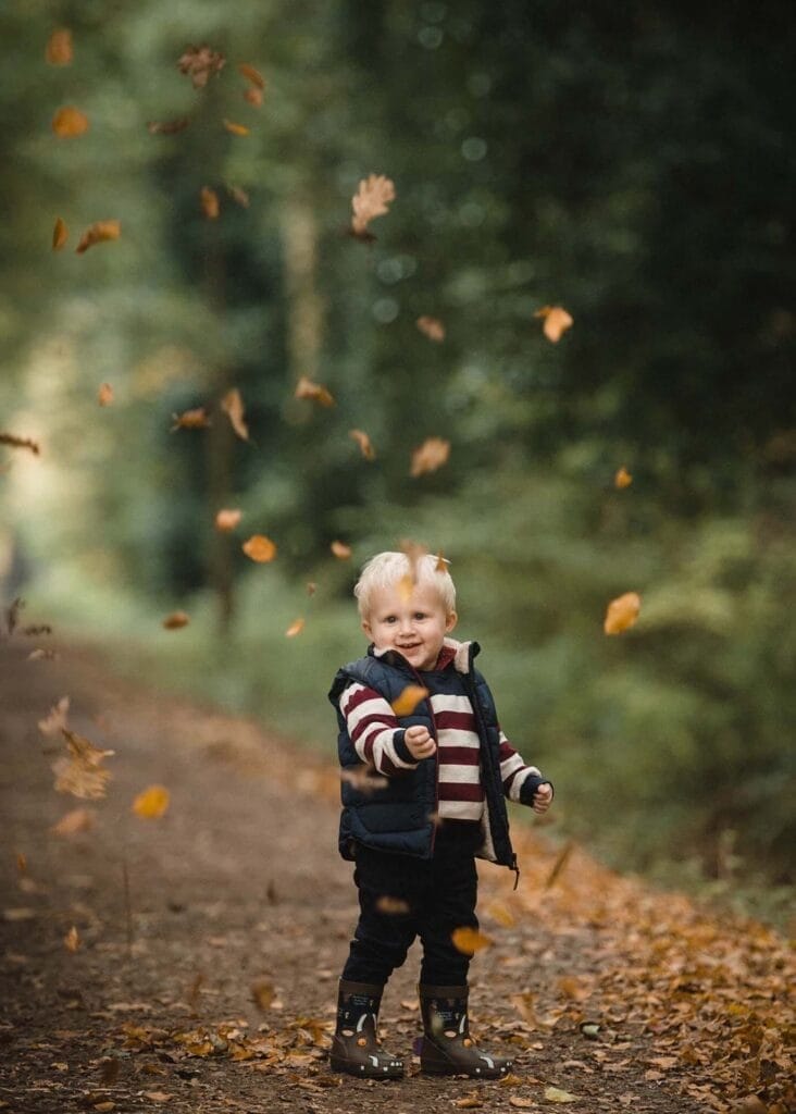 A young child in striped sweater and boots stands on a forest path, smiling and tossing autumn leaves into the air, surrounded by falling leaves and greenery.