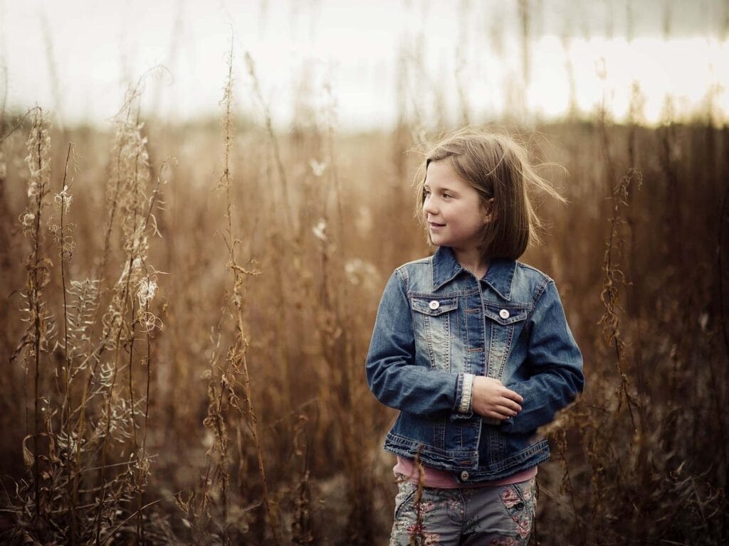 A young girl with short brown hair wearing a denim jacket stands in a field of tall, dry grass, looking to the side and smiling. The background is softly blurred, giving a warm, autumnal atmosphere.